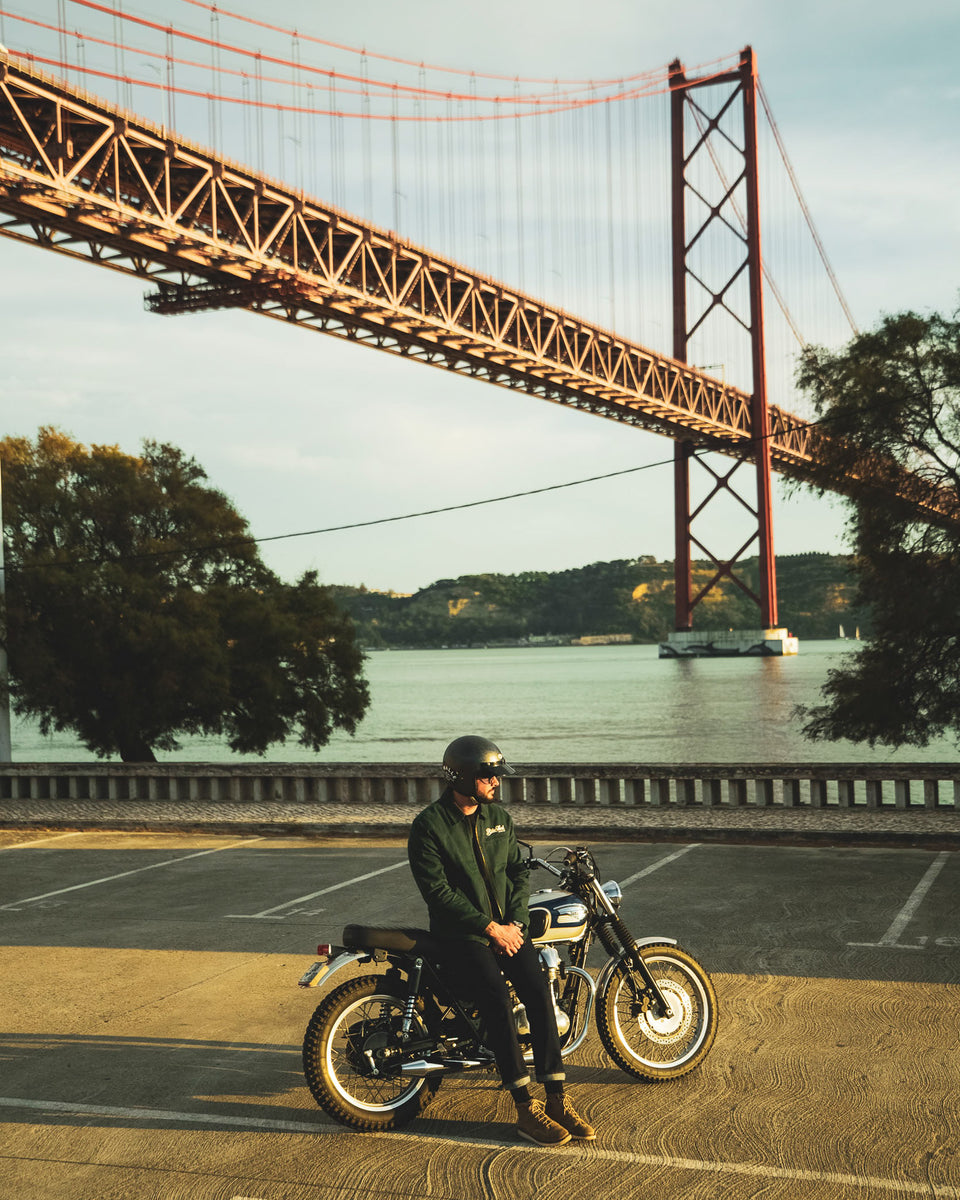 Model wearing the Bike Shed Moto Co. Twill Green logo jacket standing next to a motorcycle.