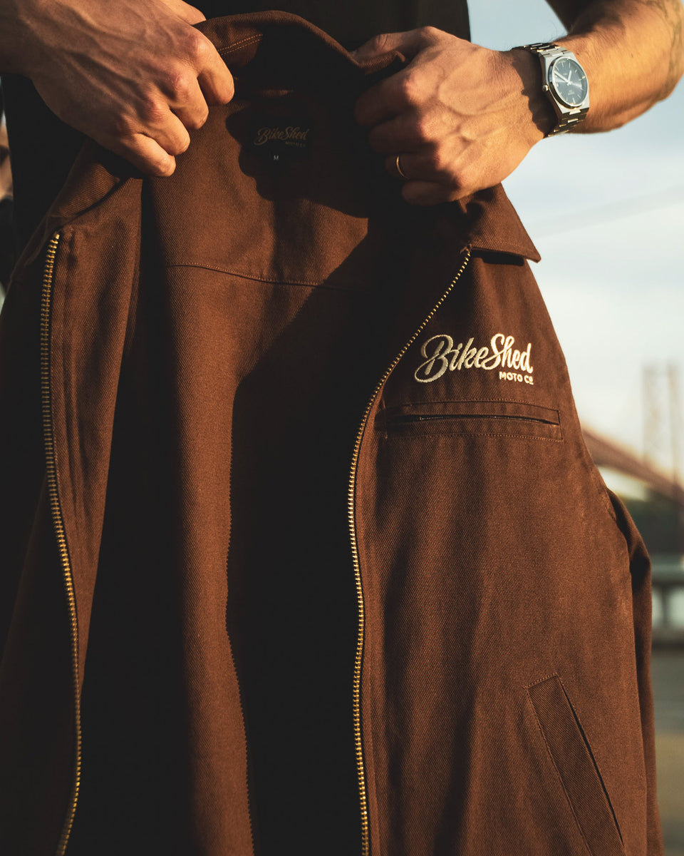 Model holding the Bike Shed Moto Co Twill brown jacket with a Bike Shed brand logo.