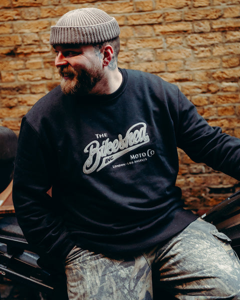 Model wearing Bike Shed Moto Co Black Applique Sweatshirt next to a motorcycle on a brick driveway
