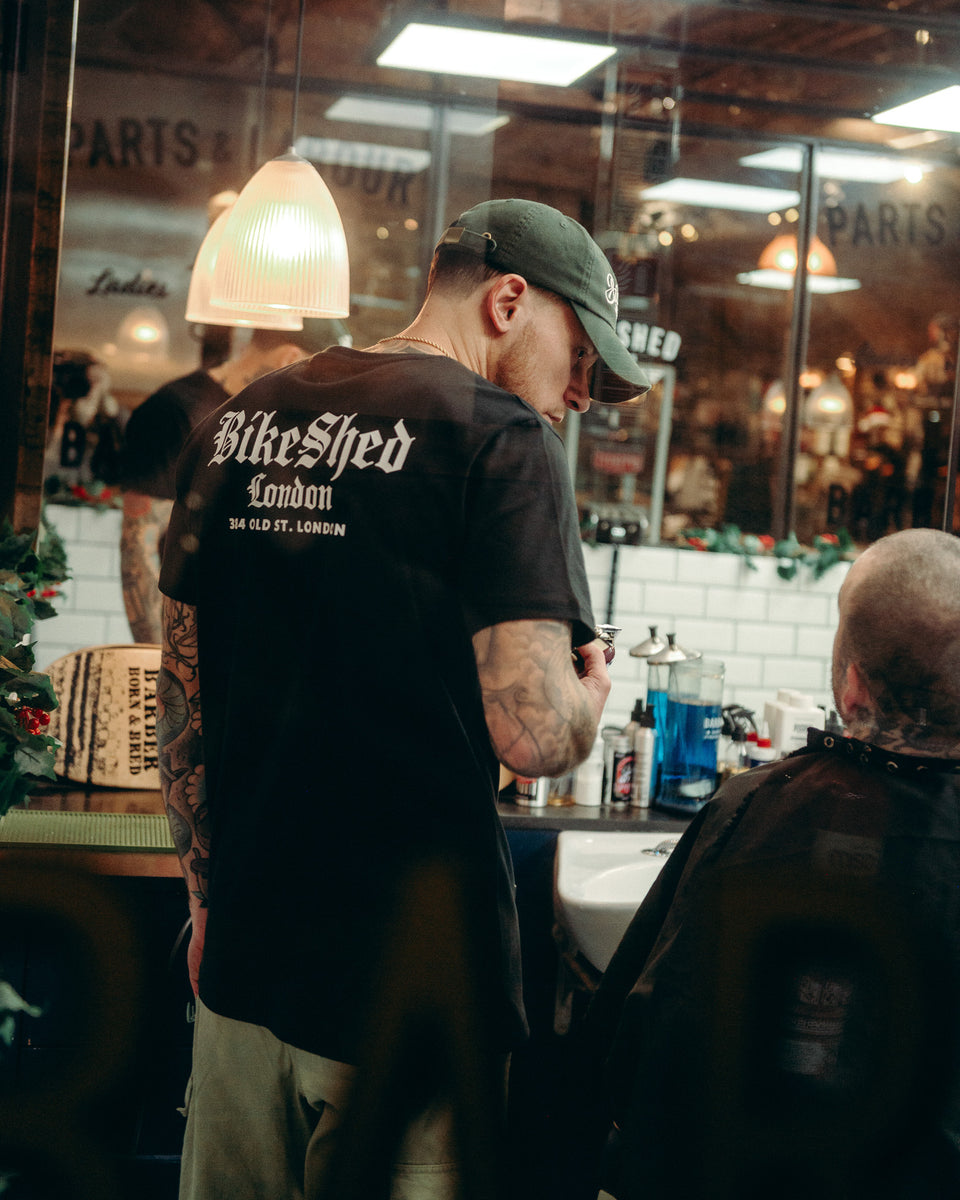 Model wearing black Bike Shed Moto Co Olde London Logo T Shirt while cutting hair in a barbershop
