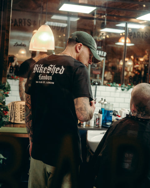 Model wearing black Bike Shed Moto Co Olde London Logo T Shirt while cutting hair in a barbershop
