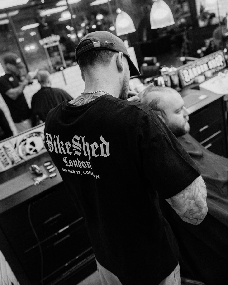 Model wearing black Bike Shed Moto Co Olde London Logo T Shirt while cutting hair in a barbershop