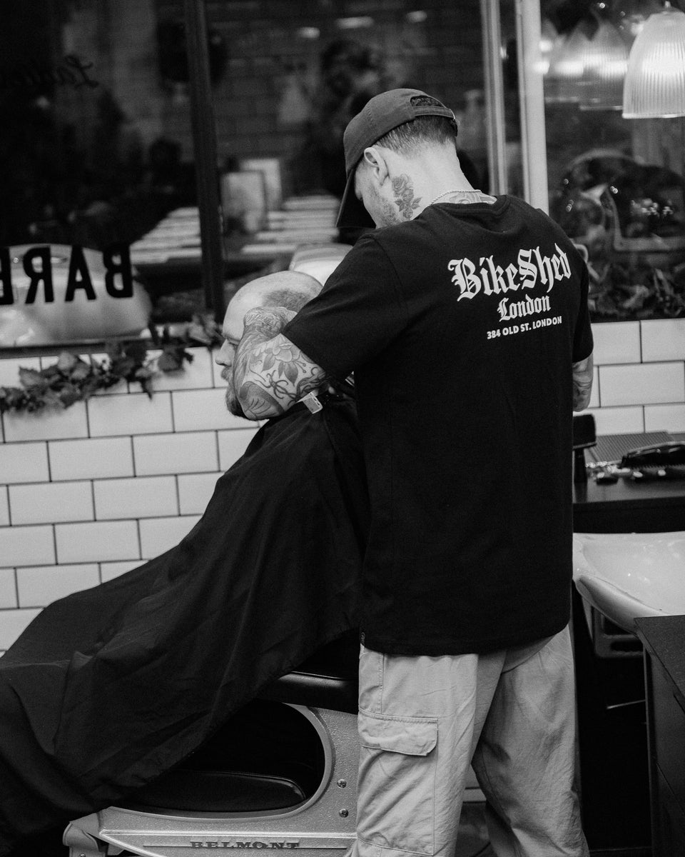 Model wearing black Bike Shed Moto Co Olde London Logo T Shirt while cutting hair in a barbershop