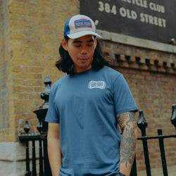 Model wearing a blue motorcycle t-shirt and cap standing in front of a brick building with a Bike Shed Moto Co sign.