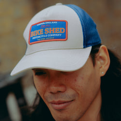 Model wearing a cream & blue motorcycle cap with 'Bike Shed Moto Co' logo against a blurred background.