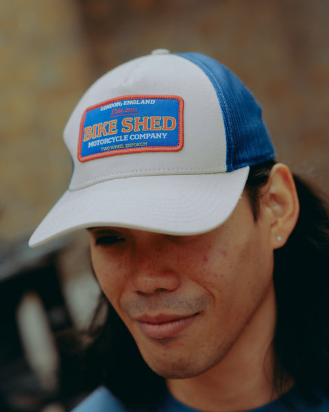 Model wearing a cream & blue motorcycle cap with 'Bike Shed Moto Co' logo against a blurred background.